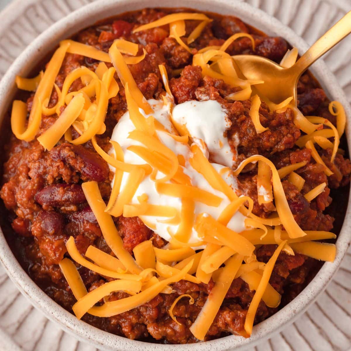 Ground beef chili topped with shredded cheddar cheese, sour cream, and a spoon in a bowl.